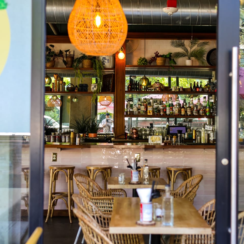 View into a restaurant with a bar, bottles, plants, and rattan chairs and tables.