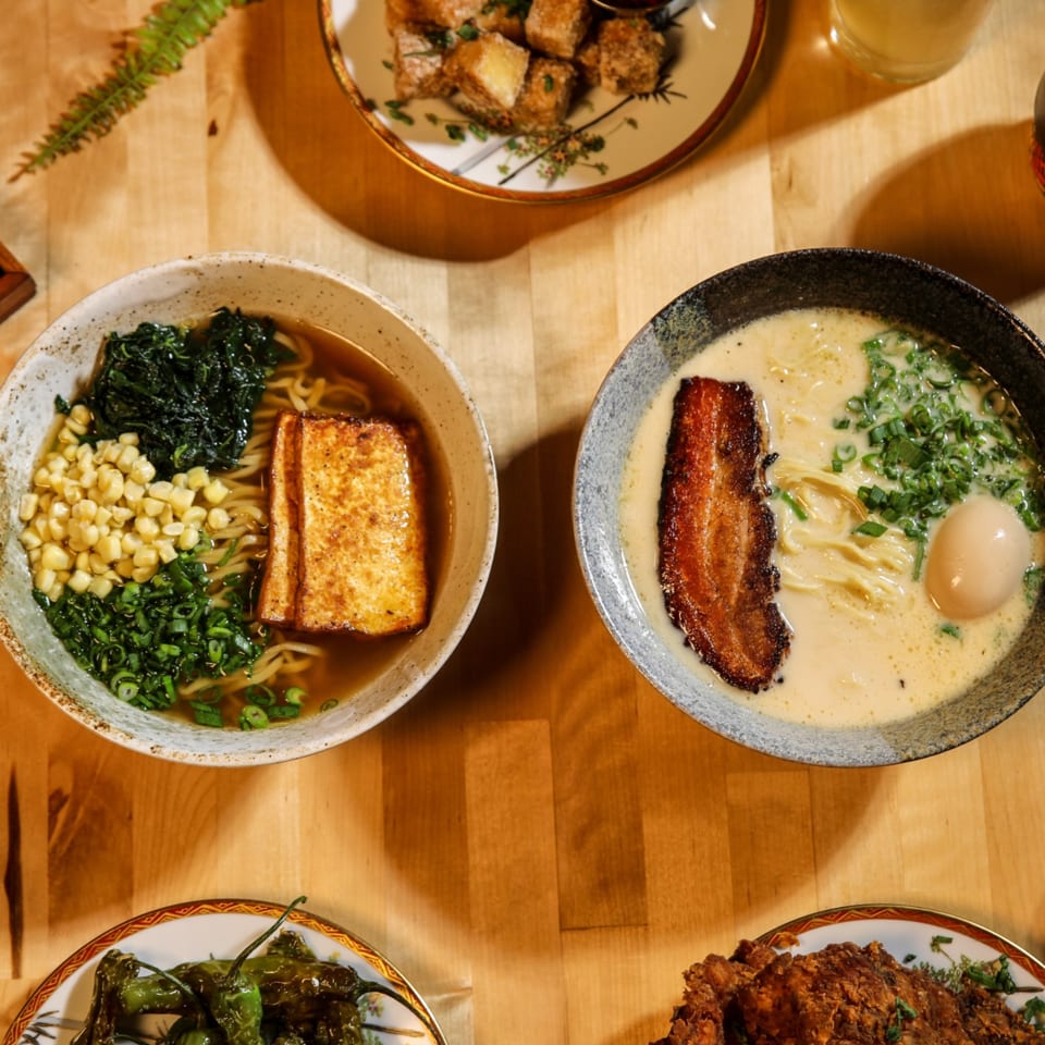 Overhead view of two ramen bowls, fried tofu, shishito peppers, and fried chicken on a wooden table.